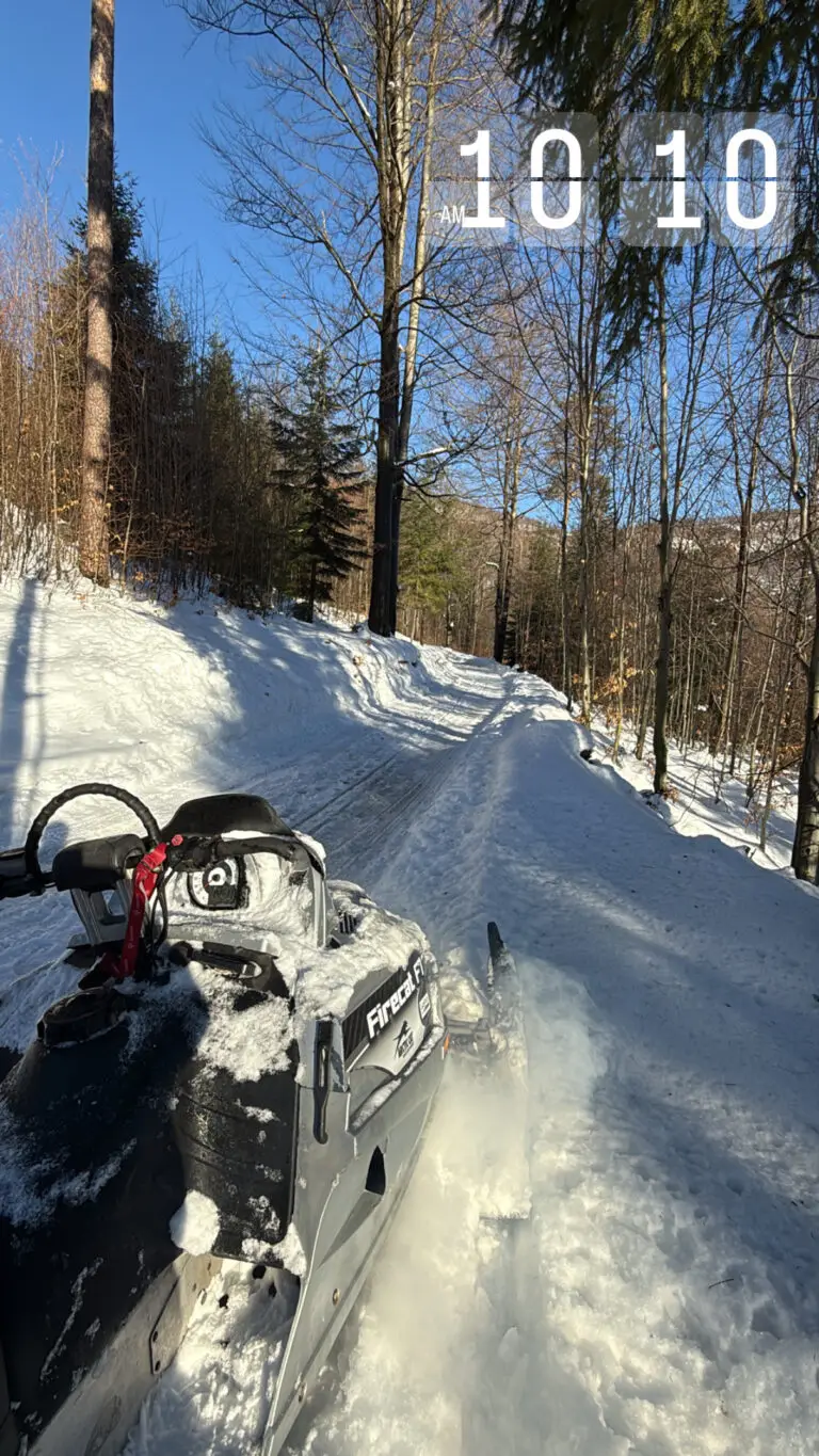 Śnieżny teren leśny z prowadzącą ścieżką, za którą widać skuter śnieżny na tle drzewa i błękitnego nieba; na drodze widoczny śnieg i parujący odcisk rury wydechowej.