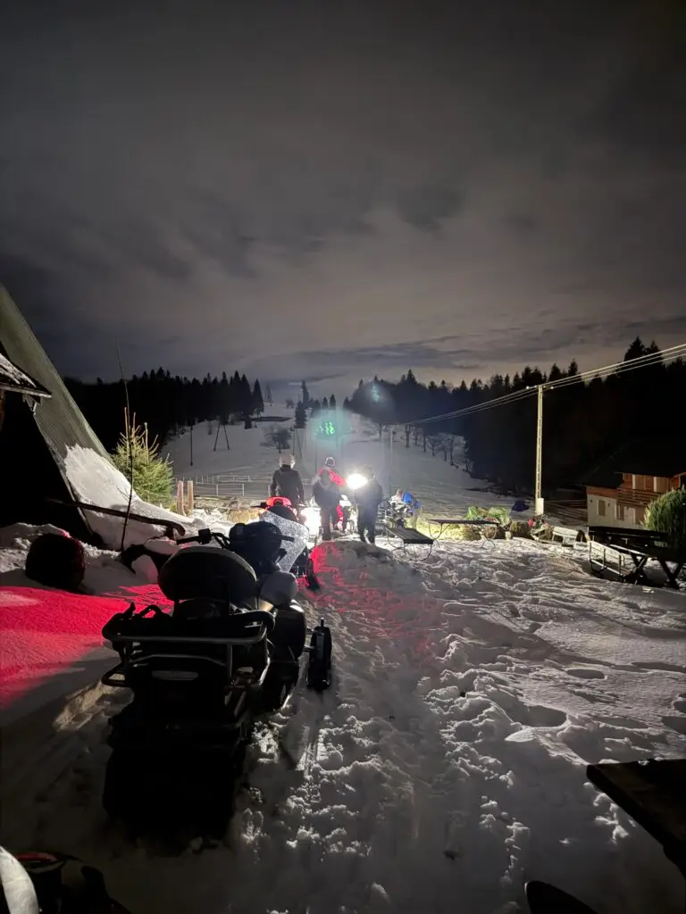 Grupa osób przy skutery śnieżne na zaśnieżonej posesji nocą, rozświetlone światłami i czerwonym światłem lasów, scena zimowa związana z jazdą na skuterach śnieżnych