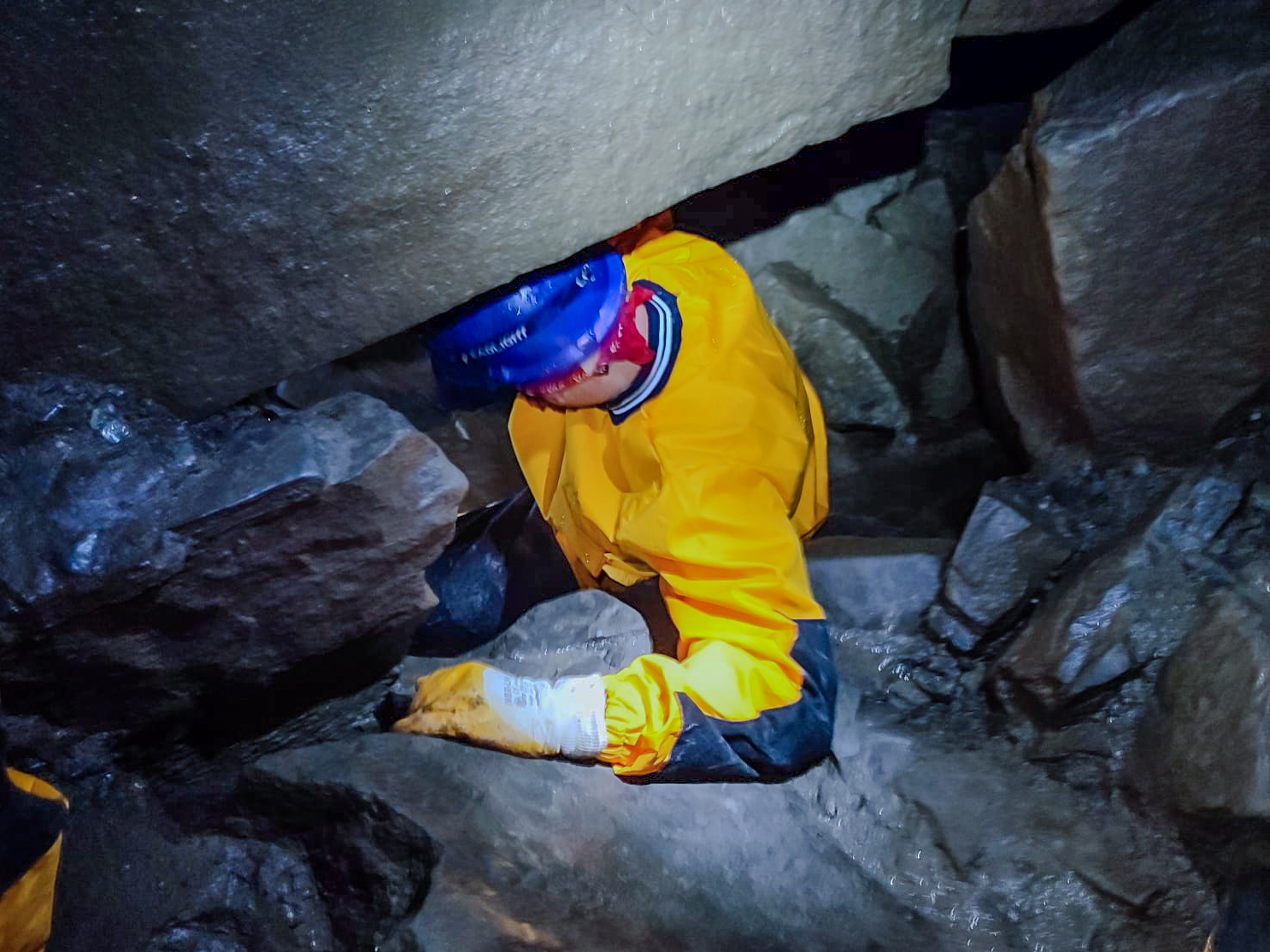 Osoba w żółtej odzieży ochronnej wspinająca się w wąskim korytarzu jaskini, z latarką na głowie.