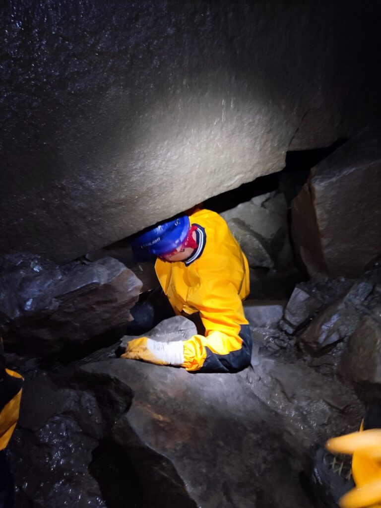 Osoba w żółtym kombinezonie wspinająca się przez szczelinę w jaskini, otoczona przez skały.