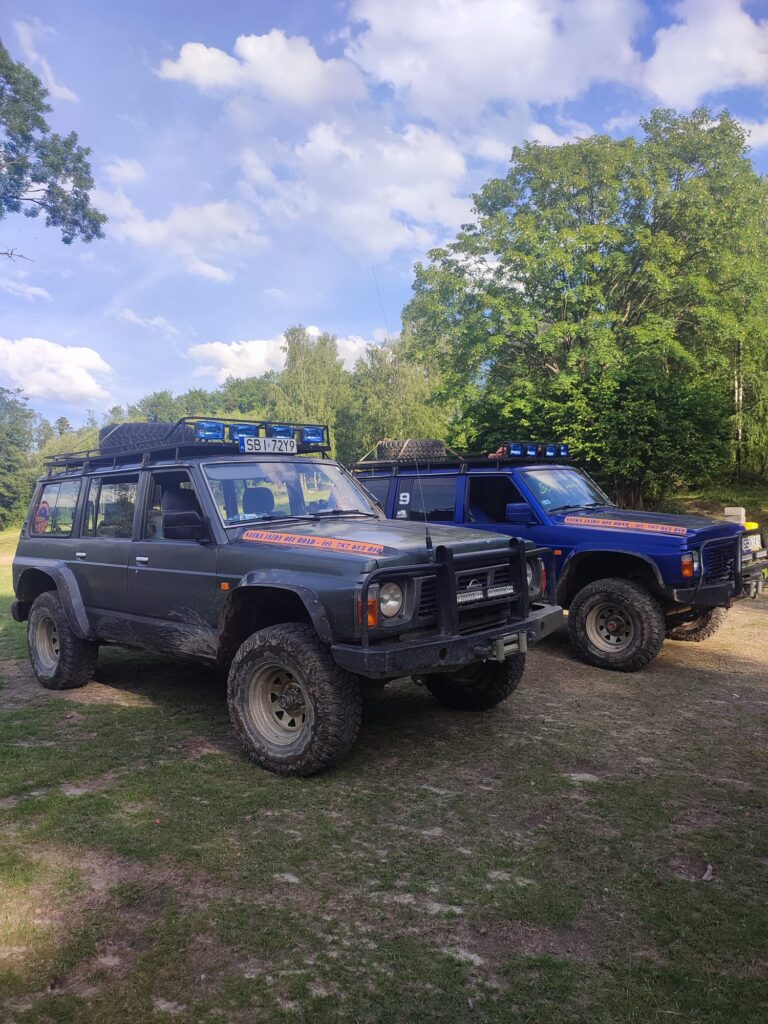 Dwa pojazdy terenowe z niebieskimi światłami na dachu, stojące na trawie w otoczeniu drzew.