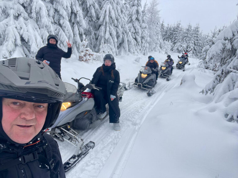 Grupa osób na skuterach śnieżnych w zimowym lesie, otoczona śniegiem i drzewami.