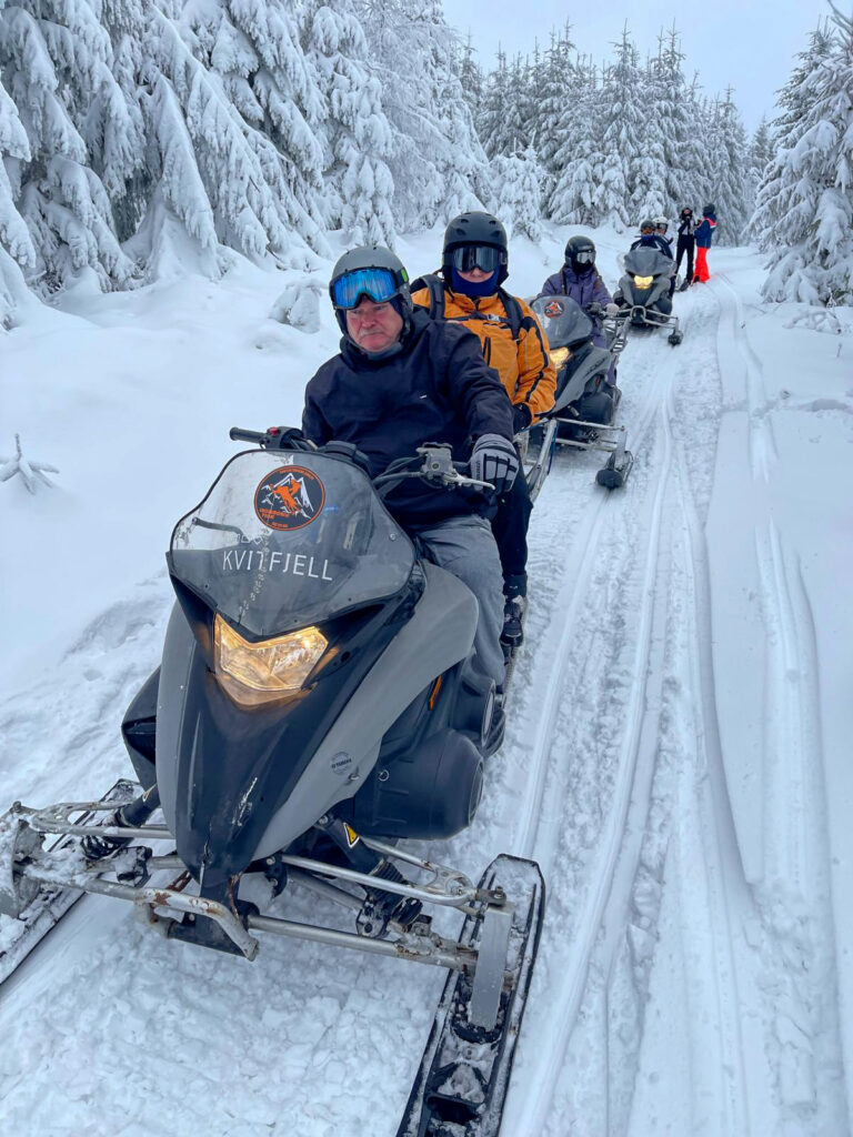 Grupa osób na skuterach śnieżnych poruszających się po ośnieżonym szlaku w lesie.