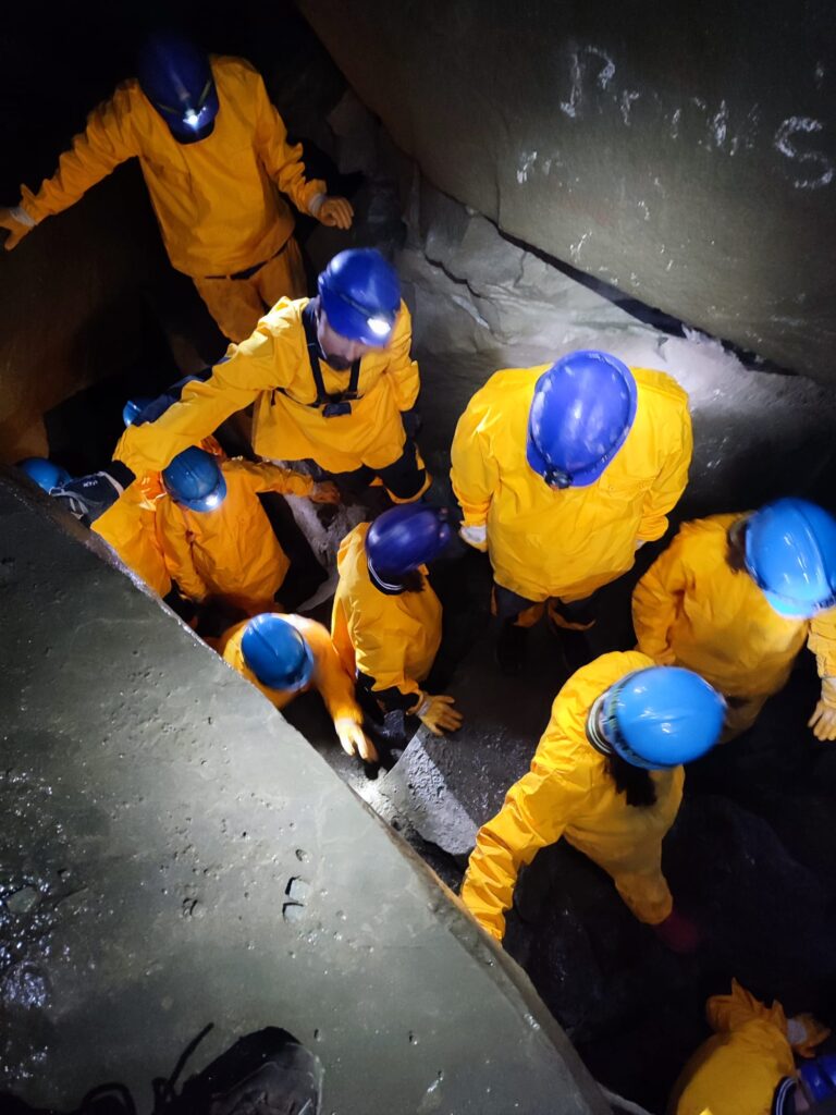 Grupa osób w żółtych kombinezonach i niebieskich kaskach schodząca w dół w wąskim korytarzu skalnym.