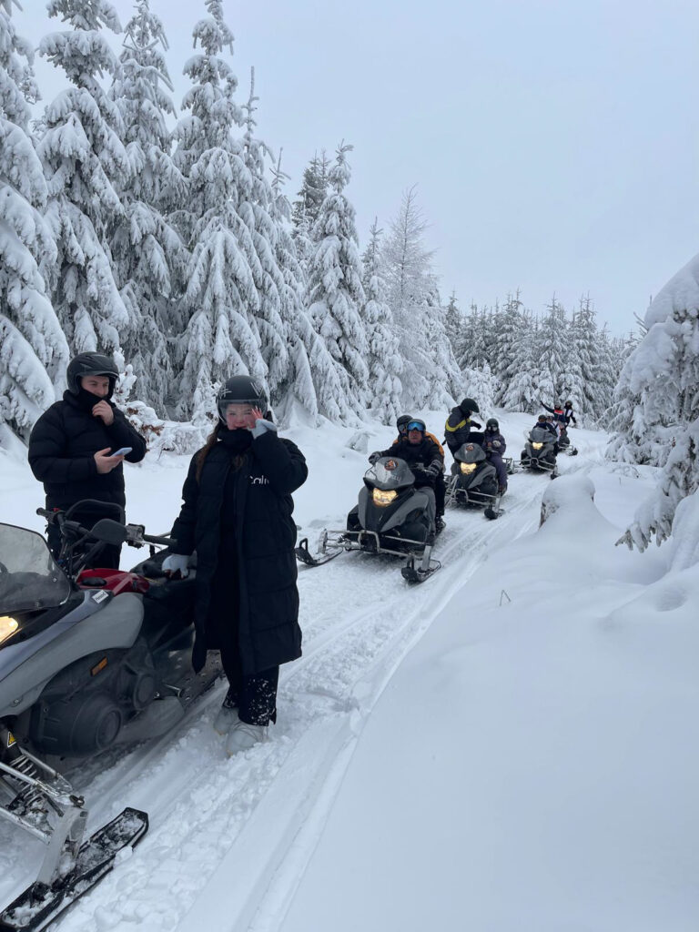 Grupa osób na skuterach śnieżnych poruszających się przez zaśnieżony las z drzewami pokrytymi śniegiem.